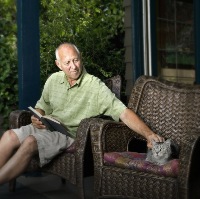 Man with metastatic pancreatic cancer sitting in outdoor chair with a book in his lap and petting a cat.